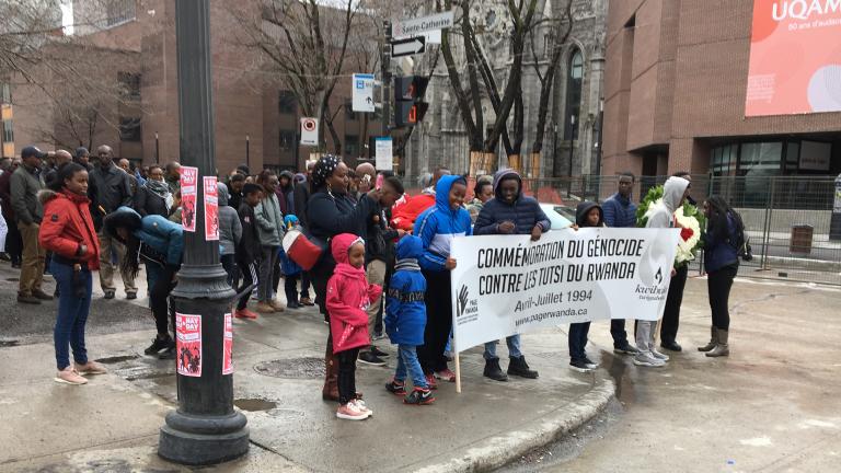 A group of people gathered on a street corner, holding a large banner reading, in French: “Commémoration du génocide contre les Tutsi du Rwanda, Avril-Juillet 1994, www.pagerwanda.ca”.