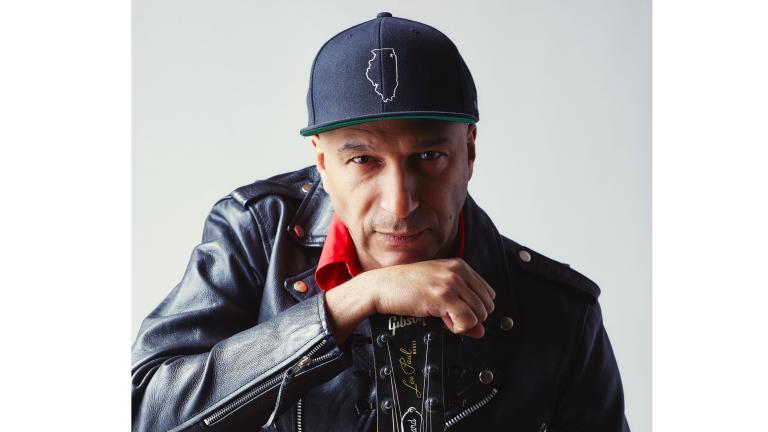 Tom Morello looks intently at the camera while leaning on his hand, which holds a guitar by the neck. He is wearing a black leather jacket, red collared shirt and baseball hat. Partially obscured.