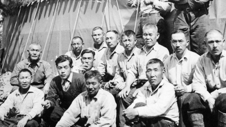 A group of men pose for a picture with a large tent and a tree in the background. Partially obscured.