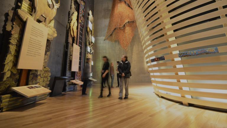 People in a Museum gallery explore tall wooden panels with wildlife and Métis beadwork. There are curved horizontal wooden slats to the right.