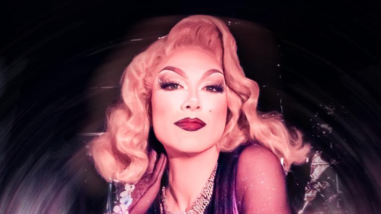 Drag queen with shoulder-length blonde hair wearing silver jewelry and a purple dress poses in front of a silver background. Partially obscured.