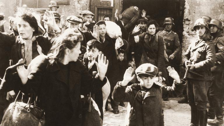 A black and white photo showing a group of people with their hands up being threatened by armed soldiers. A frightened-looking young boy is prominent in the foreground.