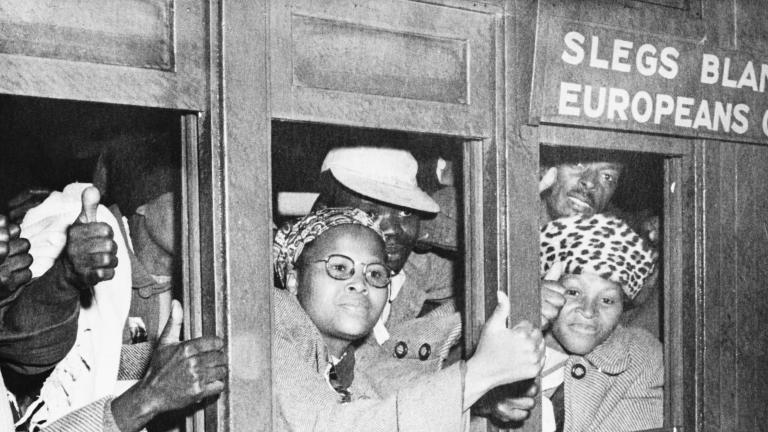 A black-and-white image of a group of black men and women look out a set of train car windows. Many are holding out their hands and making a “thumbs up” sign. A sign above the train window reads “Europeans only.”