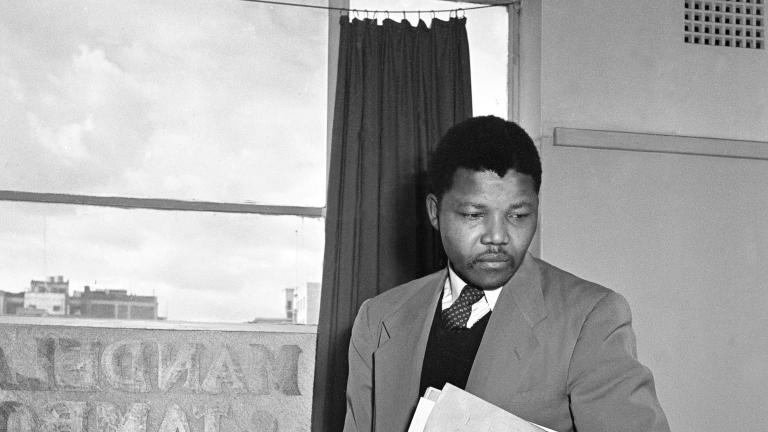 A-black-and-white image of Nelson Mandela, a young black man wearing a suit and carrying a book and some files under his left arm, standing behind a desk in an office.