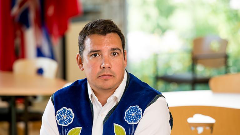 A man wearing a blue vest with flower beadwork, seated with his hands resting on a table. Partially obscured.