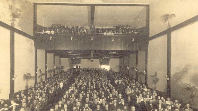  A black and white photo of a movie theatre audience. The picture is taken from the front of the theatre looking towards the back, so the faces of the audience can be seen. Potted palm trees line the walls on each side. Partially obscured.