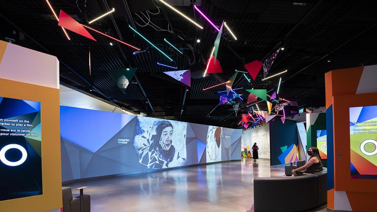 A museum gallery featuring artwork projected onto large screens and colourful geometric shapes. Partially obscured.