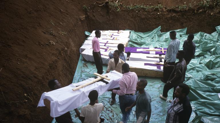 Several men carry a coffin wrapped with cloth and adorned with a large wooden cross into a large grave dug deep into the earth that already contains several similar coffins.