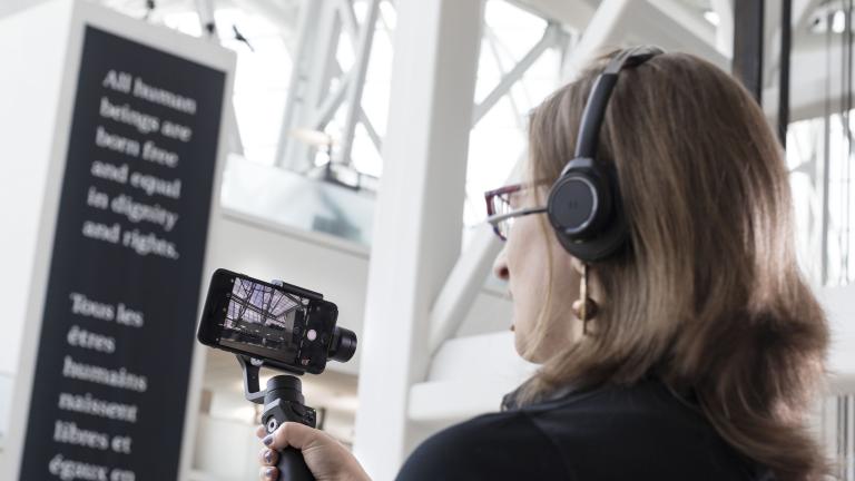 A woman holds a bar with a mobile phone attached to it and uses the screen to look at something off camera. Partially obscured.