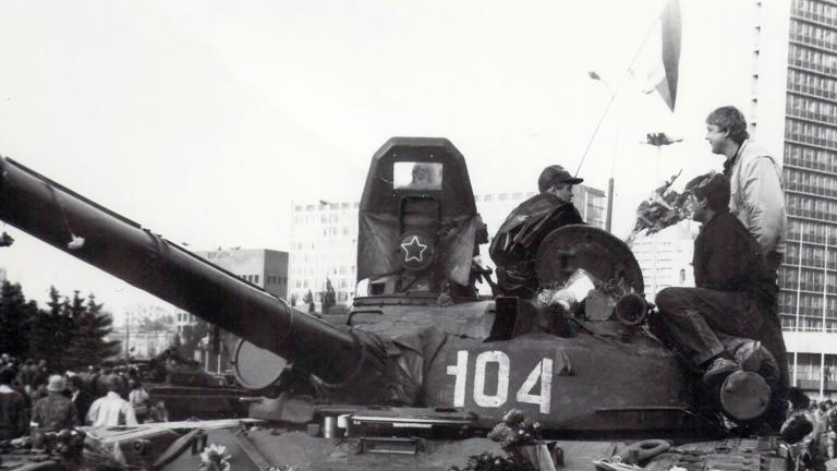 Two men stand on a parked military tank, talking. There are bouquets of flowers strewn across the tank. Behind are people milling in front of a tall building. The photo is black and white.