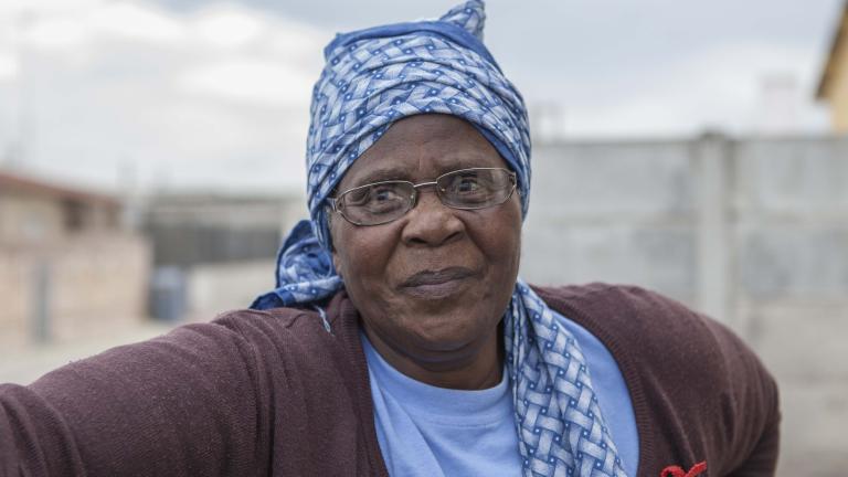 A head-and-shoulders portrait of Gogo Gladys Tyophol. She is wearing glasses and a blue patterned kerchief wrapped around her head. Her shirt says GAPA and a red AIDS ribbon is attached to her sweater. Partially obscured.