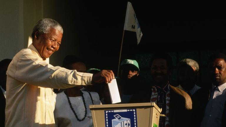 An elderly Nelson Mandela smiles as he reaches out with his right arm to put a ballot in a large metal ballot box.