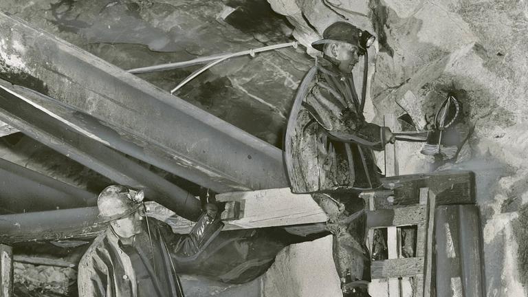 Two miners wearing helmets work underground in a uranium mine. One stands on a ladder, as the other miner watches him.