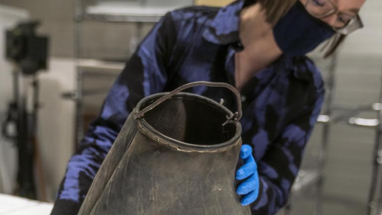 A woman holds a scuffed black rubber bag with a roughly shaped curved metal handle spanning the top.