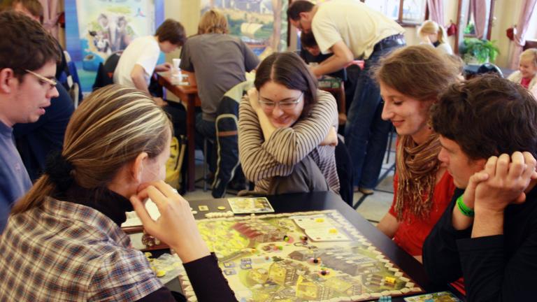 Three young women and two young men sit around a table playing a board game. There are other people playing board games at tables in the background. Partially obscured.
