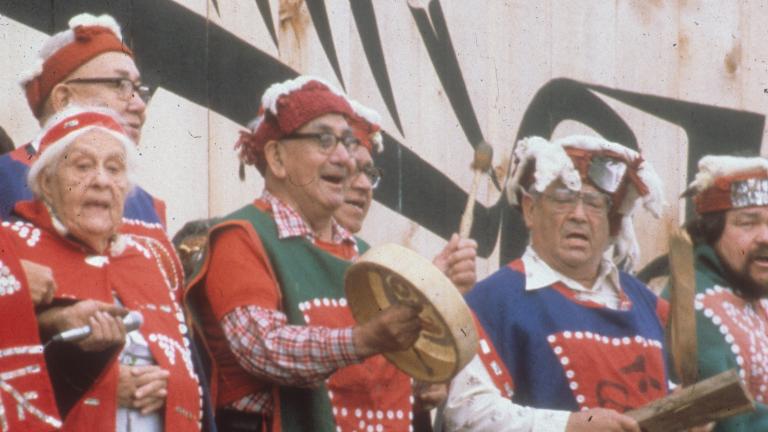 A group of older men and one woman sing while standing in front of a large wooden building. One man is playing a drum. The singers are wearing long shirts or blankets that feature patterns made from buttons.