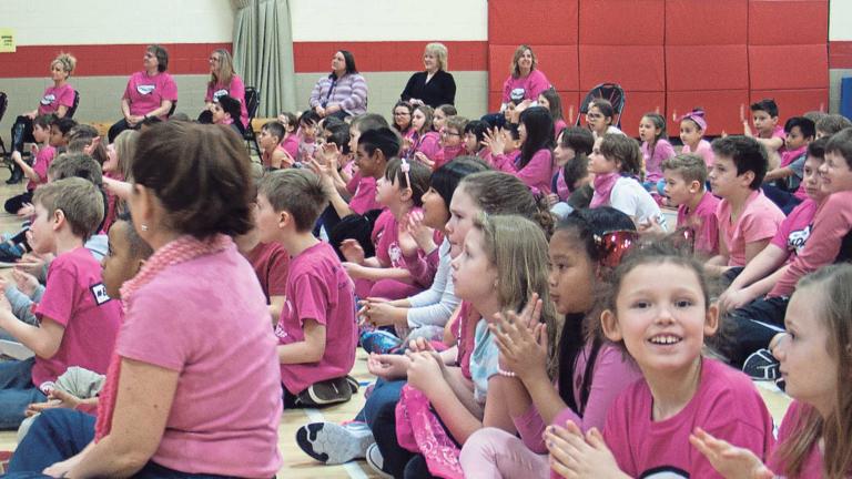 A large group of children are sitting in a school gymnasium, and all are wearing pink shirts or scarves. They are all paying attention to something out of frame, except one girl is looking directly at the camera and smiling.