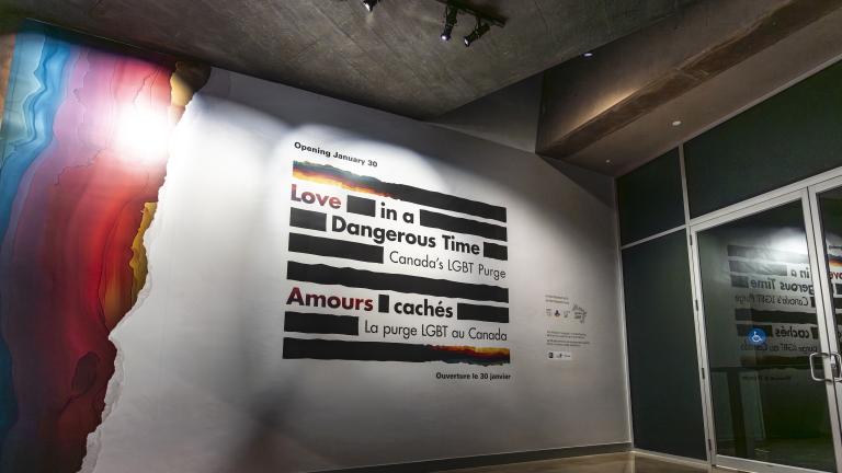 A large sign on a wall is made up of thick black horizontal lines and the English and French words “Love in a Dangerous Time: Canada’s LGBT Purge” and “Amours cachés : La purge LGBT au Canada” are distributed within the lines. The top and bottom lines are a mix of black and rainbow colours. Rainbow colours cover the left edge of the wall.