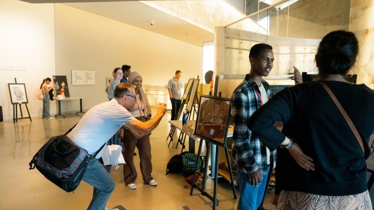 In a museum gallery, students exhibit projects on easels. Two students speak with adults about their work. Partially obscured.