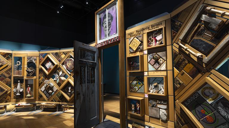 A large, curved freestanding wooden wall on which is mounted numerous small objects arranged in diamond and rectangular patterns. There is an open door in the centre of the wall. Partially obscured.