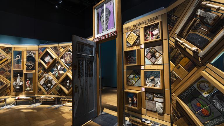 A large, curved freestanding wooden wall on which is mounted numerous small objects arranged in diamond and rectangular patterns. There is an open door in the centre of the wall. Partially obscured.