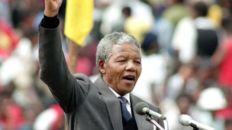 Nelson Mandela, an older dark-skinned man with short grey hair, stands in front of a podium and speaks into two microphones. He is wearing a grey suit with a tie. He is raising his right arm above his head and his hand is making a fist. Partially obscured.
