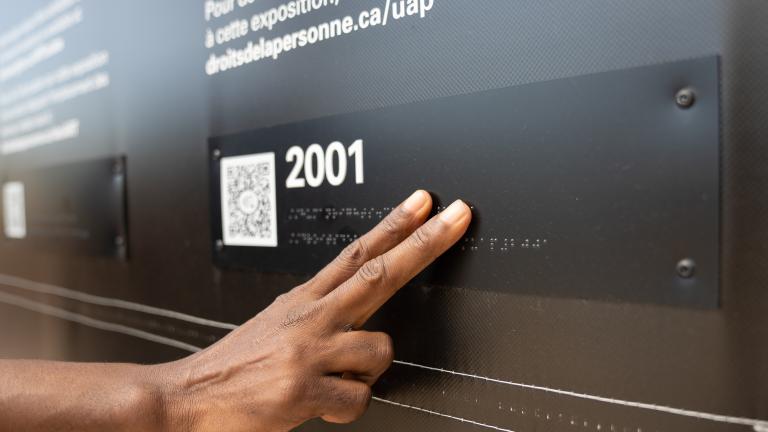 A hand touches braille letters on a museum exhibit. Partially obscured.