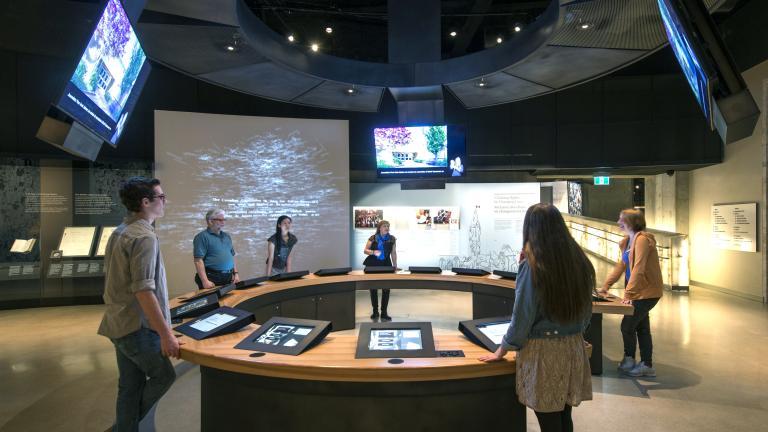 Six people in a museum gallery stand around a circular counter with video screens both embedded in the counter and hanging above them.