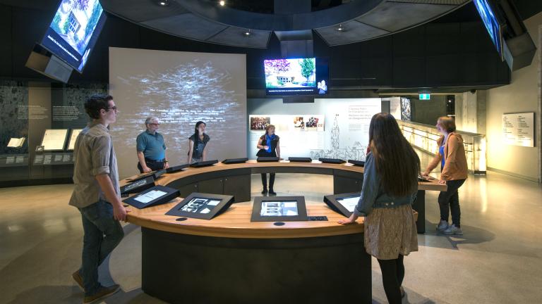 Six people in a museum gallery stand around a circular counter with video screens both embedded in the counter and hanging above them. Partially obscured.