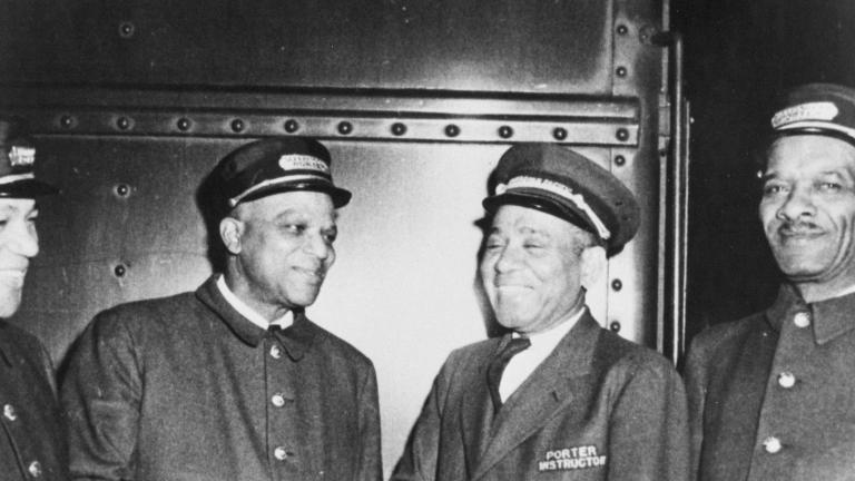 A black and white photo of four men in train porter uniforms. All of the men are smiling, and the two men in the middle appear to be shaking hands.