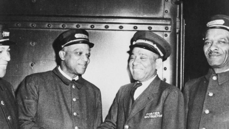 A black and white photo of four men in train porter uniforms. All of the men are smiling, and the two men in the middle appear to be shaking hands. Partially obscured.