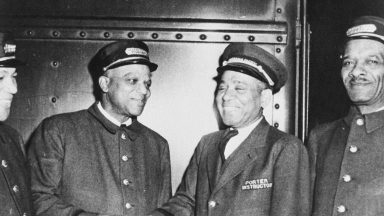 A black and white photo of four men in train porter uniforms. All of the men are smiling, and the two men in the middle appear to be shaking hands. Partially obscured.