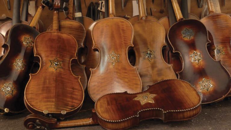 Several restored violins are pictured leaning on each other in a pile. Each of the violins carry at least one Star of David Partially obscured.