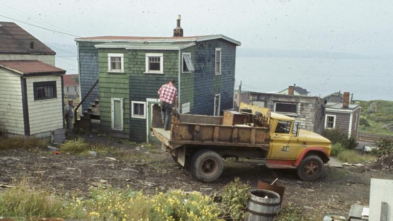 A man stands on the back of a garbage truck that is parked in front of a wooden house with green shingling. In the background rail lines and more houses can be seen, and behind that, Halifax harbor.