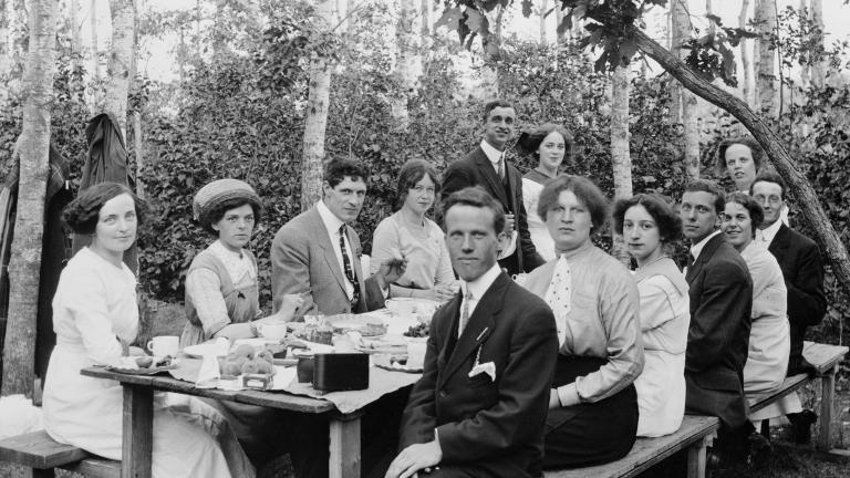A group of well-dressed men and women sitting at a picnic table with trees in the background.