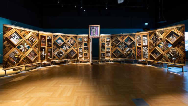 A large, curved freestanding wooden wall on which is mounted numerous small objects arranged in diamond and rectangular patterns. There is an open door in the centre of the wall. Partially obscured.