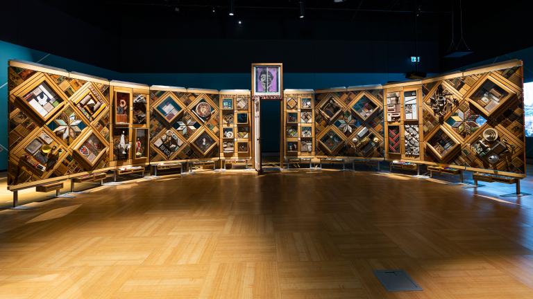 A large, curved freestanding wooden wall on which is mounted numerous small objects arranged in diamond and rectangular patterns. There is an open door in the centre of the wall. Partially obscured.