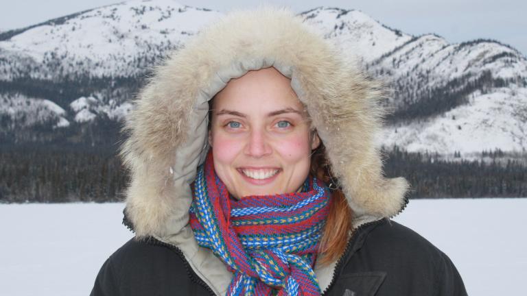 Smiling female wearing a fur-lined parka posing in front of a mountainous terrain. Partially obscured.