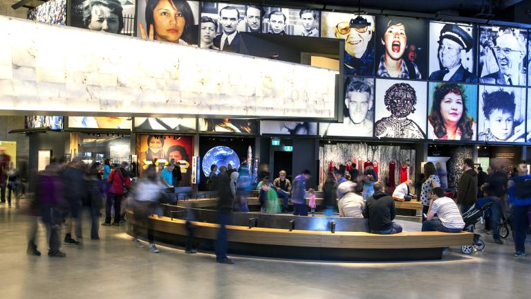 A large crowd in a museum gallery. There is a curved bench in the centre of the room. The display consists of a series of portraits above alcoves each containing a different exhibit.