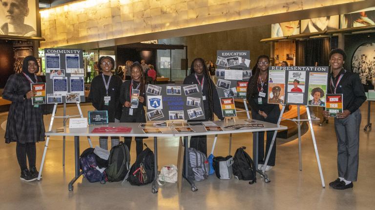 Six black students in a museum hall present proudly their work on posters and on a table. Partially obscured.