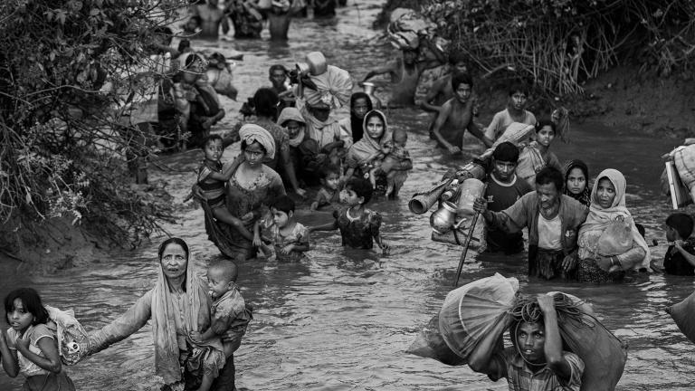 Rohingya women, children and men wade through waist-high muddy river water. Some carry young children, while others carry bags of possessions, including household items. Partially obscured.