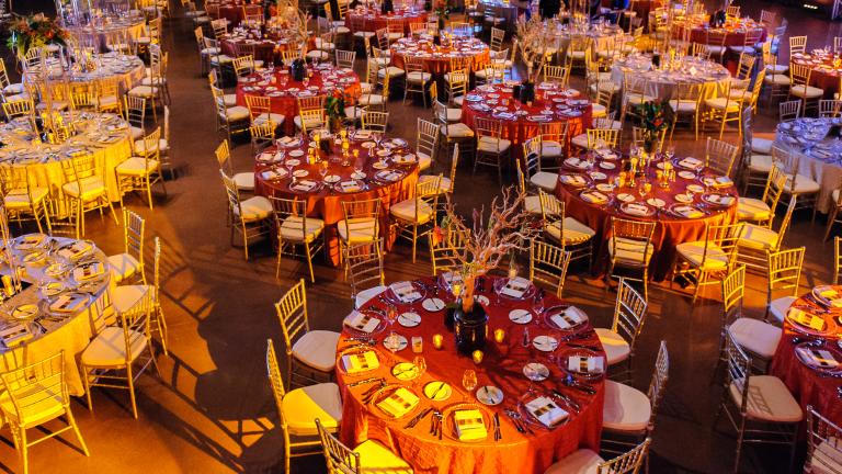 Several round tables, each with formal table settings for 10 guests, viewed from above. An orange glow is cast upon the scene.