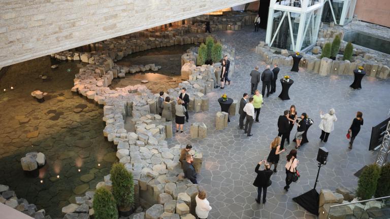 A crowd of people mingling in an area surrounded by light brown or grey stones, small pools of clear water and a small bit of green foliage.