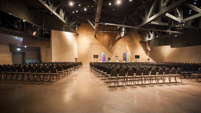 A large room with high ceilings and a polished brown concrete floor. A few hundred chairs are lined up in neat rows facing a stage.