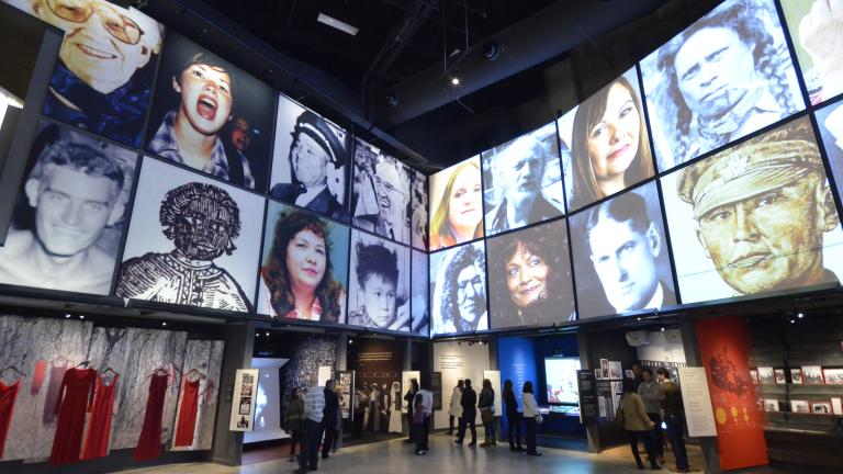 People explore a Museum gallery with two rows of square portraits above a series of alcoves, containing photographs, videos, and text. Partially obscured.