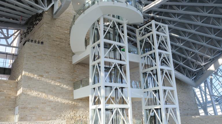 The interior of a large open spaced part of the Museum with two elevator shafts in the centre of the image, and a limestone wall.