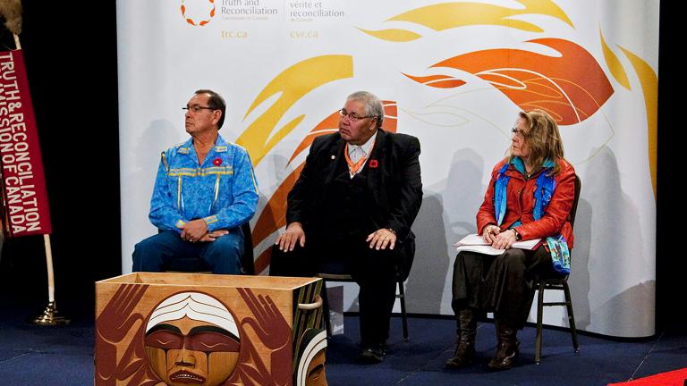 Two men and a woman sit in front of a white background decorated with stylized flames. To their right is a vertical banner reading "Truth & Reconciliation Commission of Canada", and in front of them is a rectangular carved wooden box. On the side facing the camera is a round, weeping face.