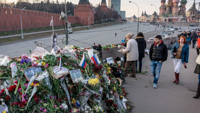A huge pile of flowers, photographs and messages sits on a bridge beside a road. People are walking over the bridge. Some are looking at the pile of mementos. One woman is taking a photograph with her tablet. In the background is a red concrete wall (Red Square in Moscow) and a church with curvy, domed spires (St. Basil’s Cathedral).