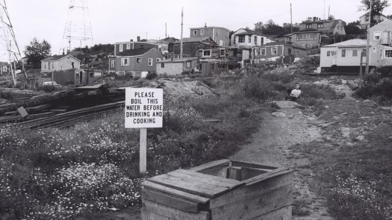 A black and white image of about 15 houses on a hill. In the foreground is a well with a sign beside it which reads: “Please boil this water before drinking and cooking.