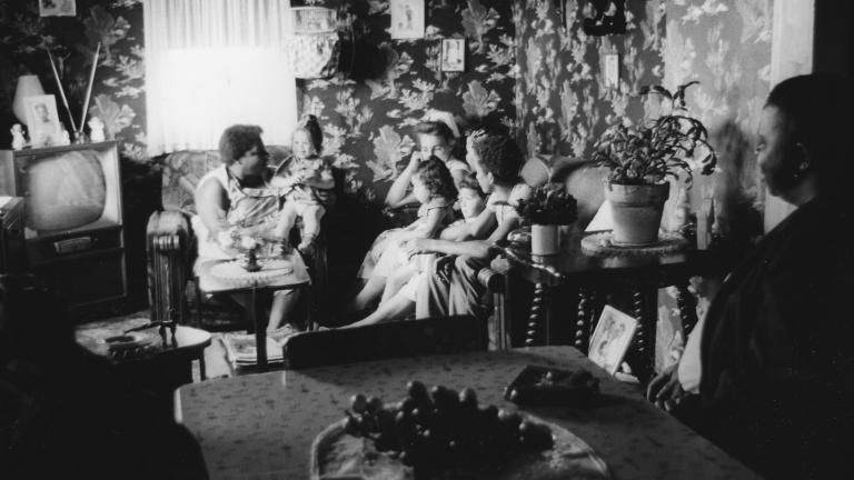 A black and white image of women and young children sitting on couches and chairs in a small living room.
